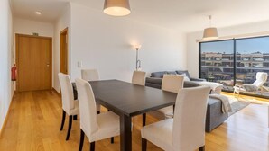 A modern living room with a dining table, chairs, a sofa, and a large window with a view of buildings. - Jardins do Oceano in Cani o (Santa Cruz)