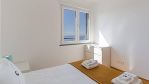 A hotel room with a bed, a wooden bedside table, a window with a view of the sea, and a white wall. - Jardins do Oceano in Cani o (Santa Cruz)