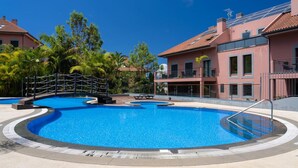A swimming pool with a surrounding deck, palm trees, and a building with a red roof. - Santa Luzia Funchal View 2 Bedrooms Pool (Funchal)