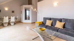 A modern living room with a glass coffee table, a grey sofa, and a dining area with white chairs. - Santa Luzia Funchal View 2 Bedrooms Pool (Funchal)