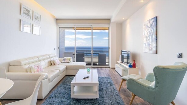 Una sala de estar moderna con un sofá, una mesa de centro y un televisor. Hay un balcón con vista al mar.