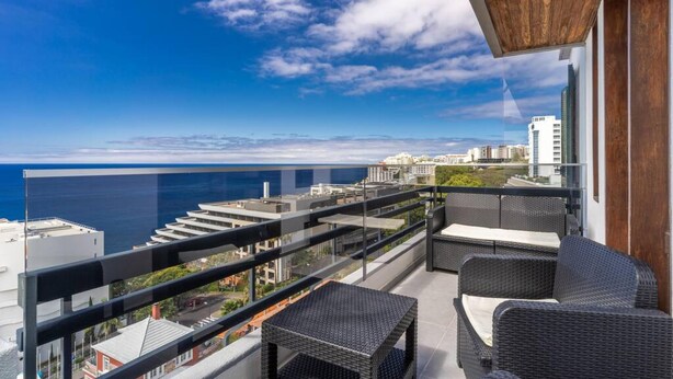 A balcony with wicker furniture, a glass railing, and a view of the ocean and cityscape. - Monumental Apartment 712 Balcony Sea View (Funchal)