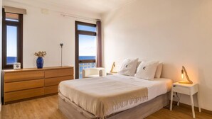 A bedroom with a large bed, wooden dresser, bedside tables, and a view of the sea through a window. - Monumental Apartment 712 Balcony Sea View (Funchal)