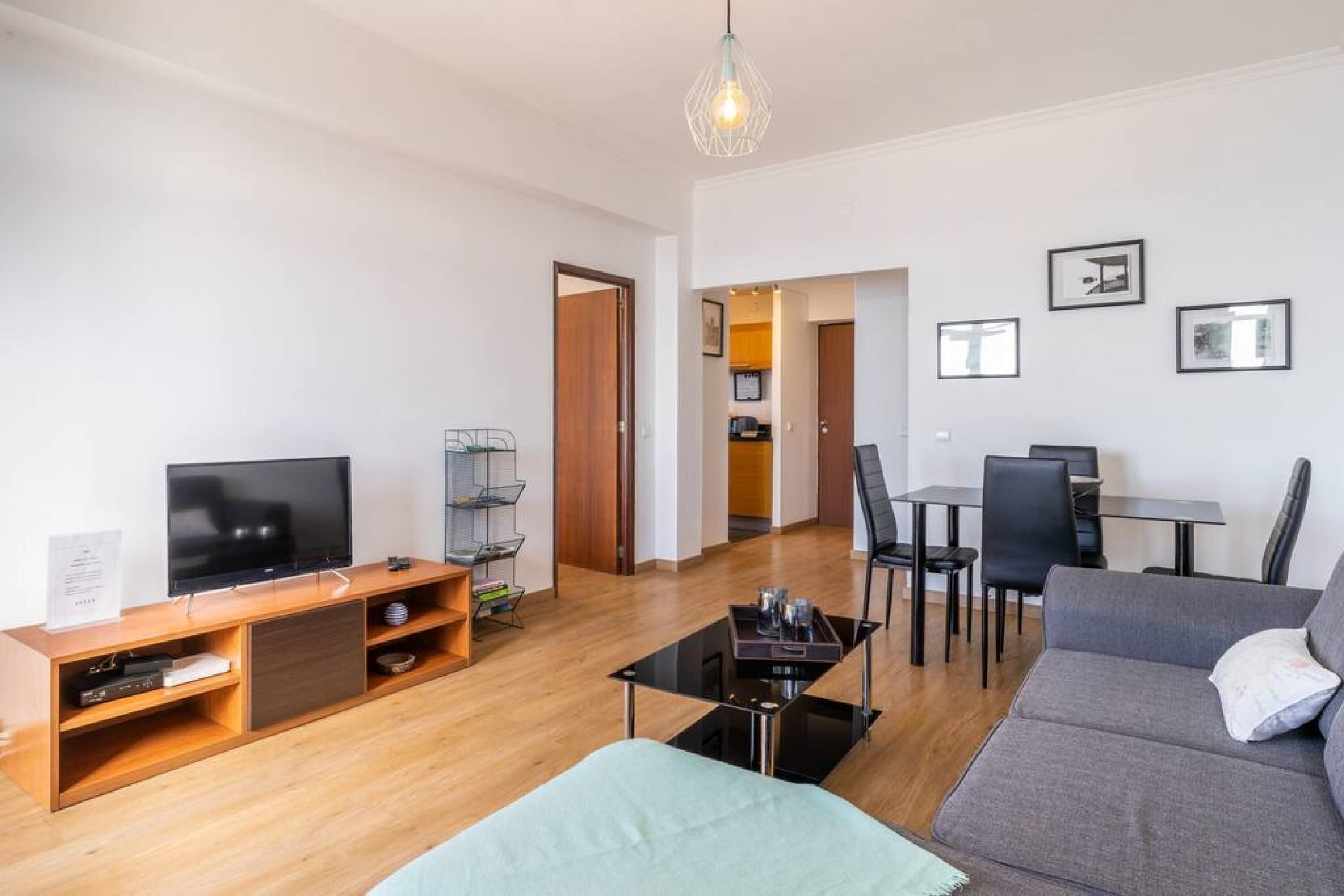 A modern living room with a flat-screen TV, wooden entertainment unit, and a sofa. There's a dining area with a table and chairs, and a kitchen visible through an open doorway.