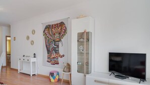 A modern living room with a large colorful elephant painting, a white cabinet with glass doors, and a flat-screen TV. - Alta do Garajau II 2 AT (Santa Cruz)