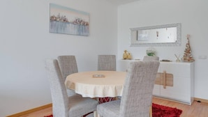 A dining area with a round table, four grey upholstered chairs, a red rug, a white cabinet with decorative items, and a cityscape painting on the wall. - Alta do Garajau II 2 AT (Santa Cruz)