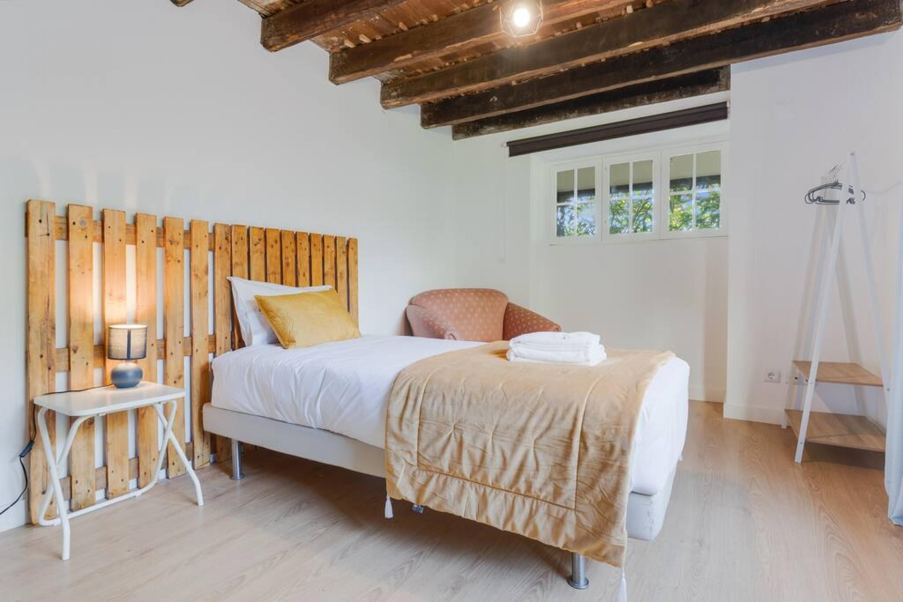 A bedroom with a wooden headboard, a bed with white bedding, a bedside table with a lamp, and a window with a view of greenery.