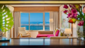 Une chambre d’hôtel avec une grande fenêtre, un canapé, un fauteuil, une lampe et un vase rempli de fleurs.