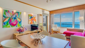 Une chambre d’hôtel moderne dotée d’un coin repas, d’une télévision, d’une grande fenêtre donnant sur la mer et de deux tableaux colorés accrochés au mur.