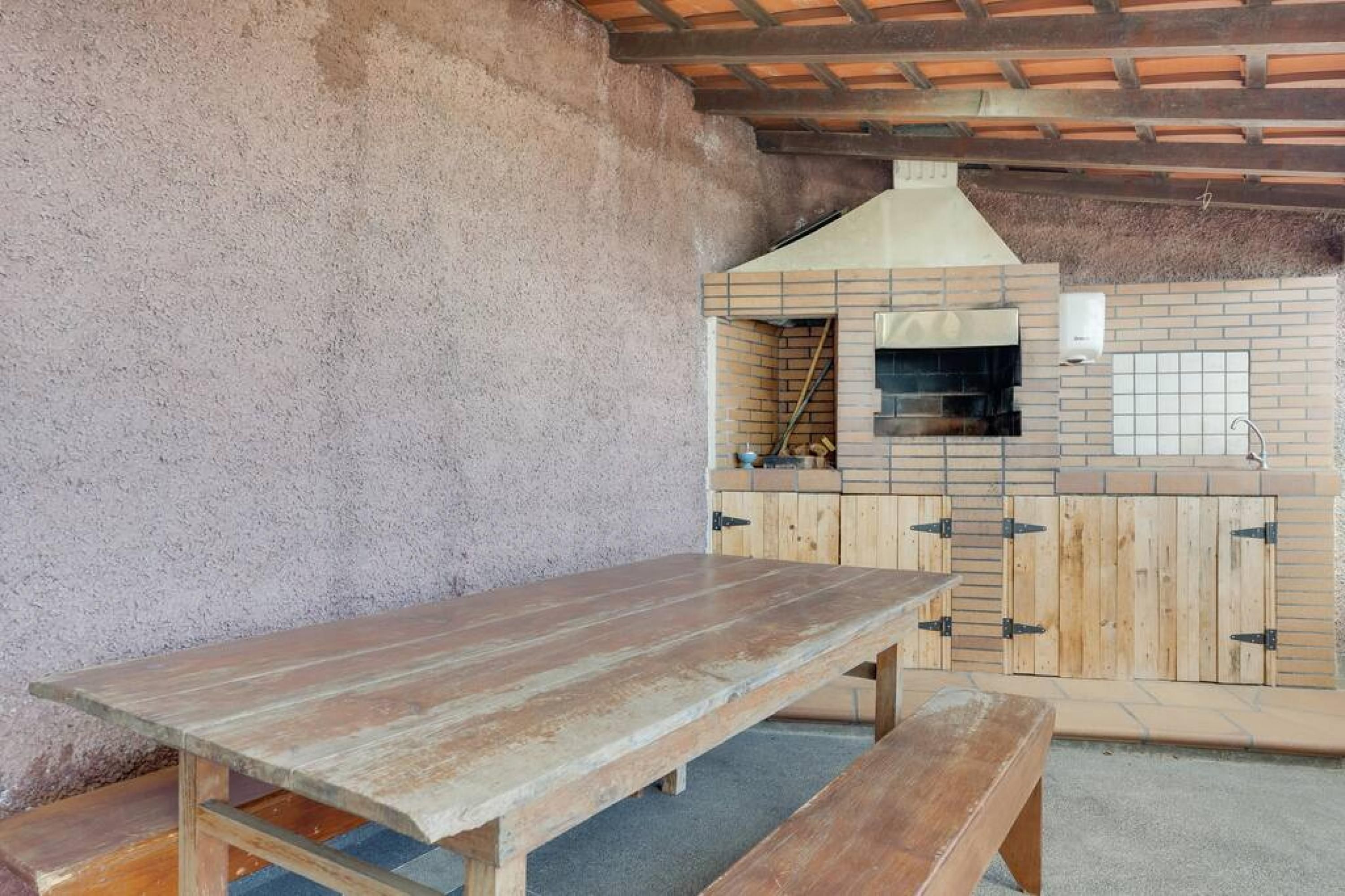 An outdoor patio with a wooden table and benches, a brick oven, and a tiled backsplash.