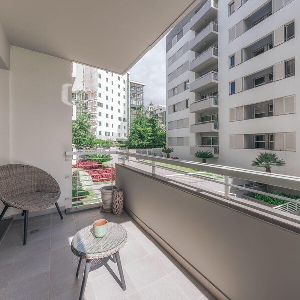 A balcony with a wicker chair and a small table, overlooking a garden and adjacent buildings.