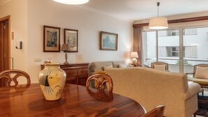 Een eetgedeelte met een houten tafel, stoelen en een vaas. Achtergrond: een bank en een raam met gordijnen.