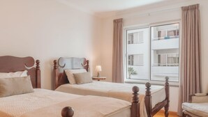Hotelkamer met twee houten bedden, een raam met gordijnen, een klein tafeltje met een lamp en uitzicht op een gebouw.