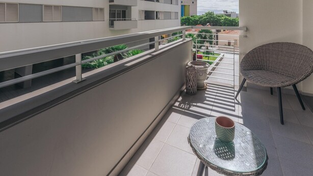 Een balkon met een glazen tafel en een rieten stoel, met uitzicht op een woongebied en een gebouw op de achtergrond.