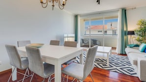 A modern living room with a dining area, large windows, and a view of buildings outside. - Ajuda I 4 D in Funchal (Funchal)