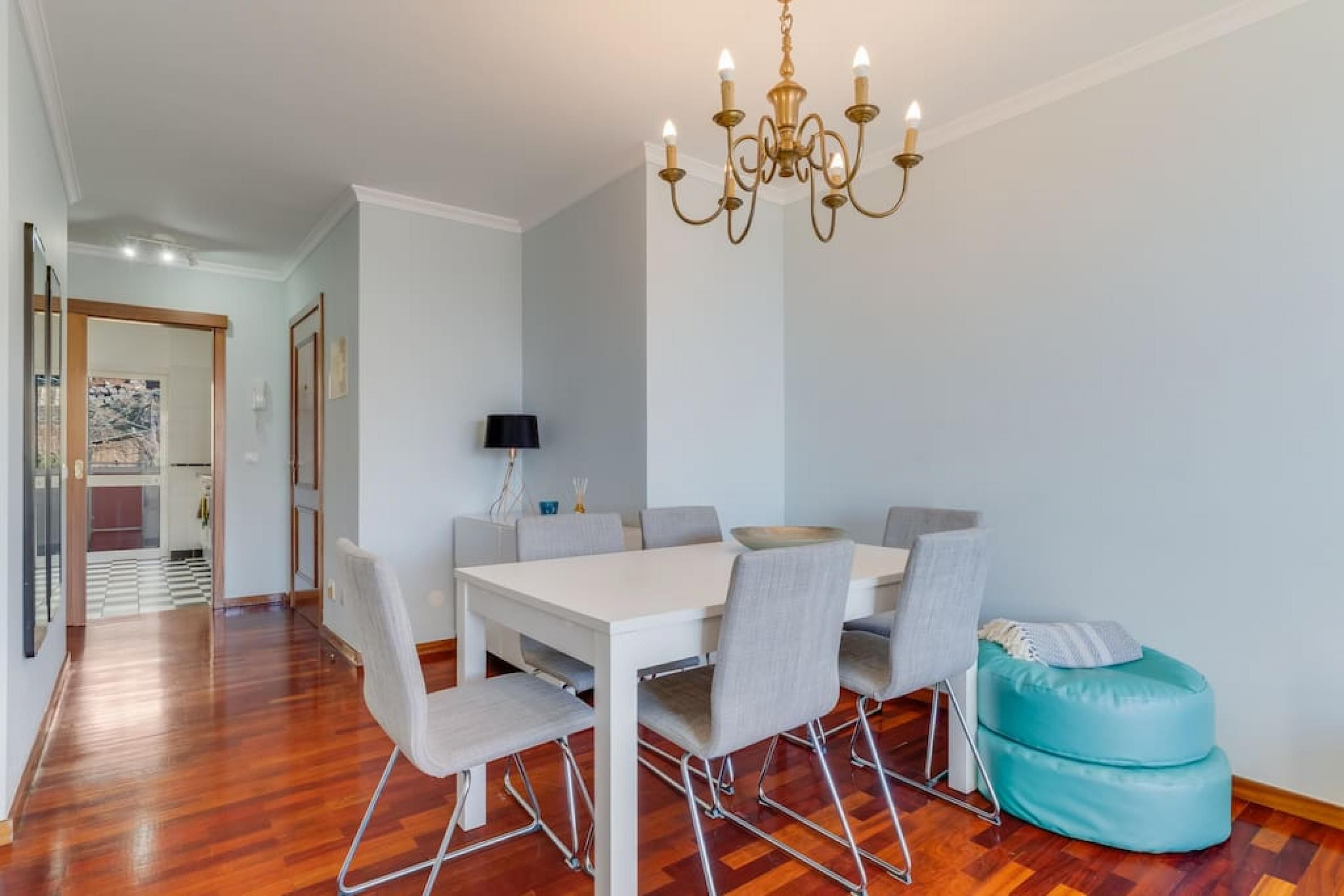 Een eetgedeelte met een witte tafel, grijze stoelen, een turquoise poef en een kroonluchter.