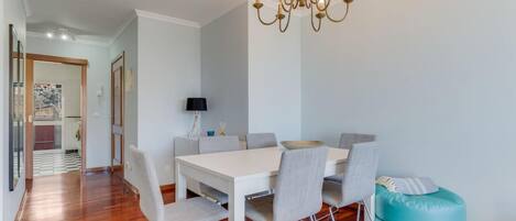 Un área de comedor con una mesa blanca, sillas grises, un otoman turquesa y una araña.