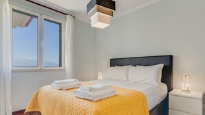 A neatly made bed with a yellow bedspread and white pillows, a bedside table with a lamp, and a window with curtains. - Ajuda I 4 D in Funchal (Funchal)