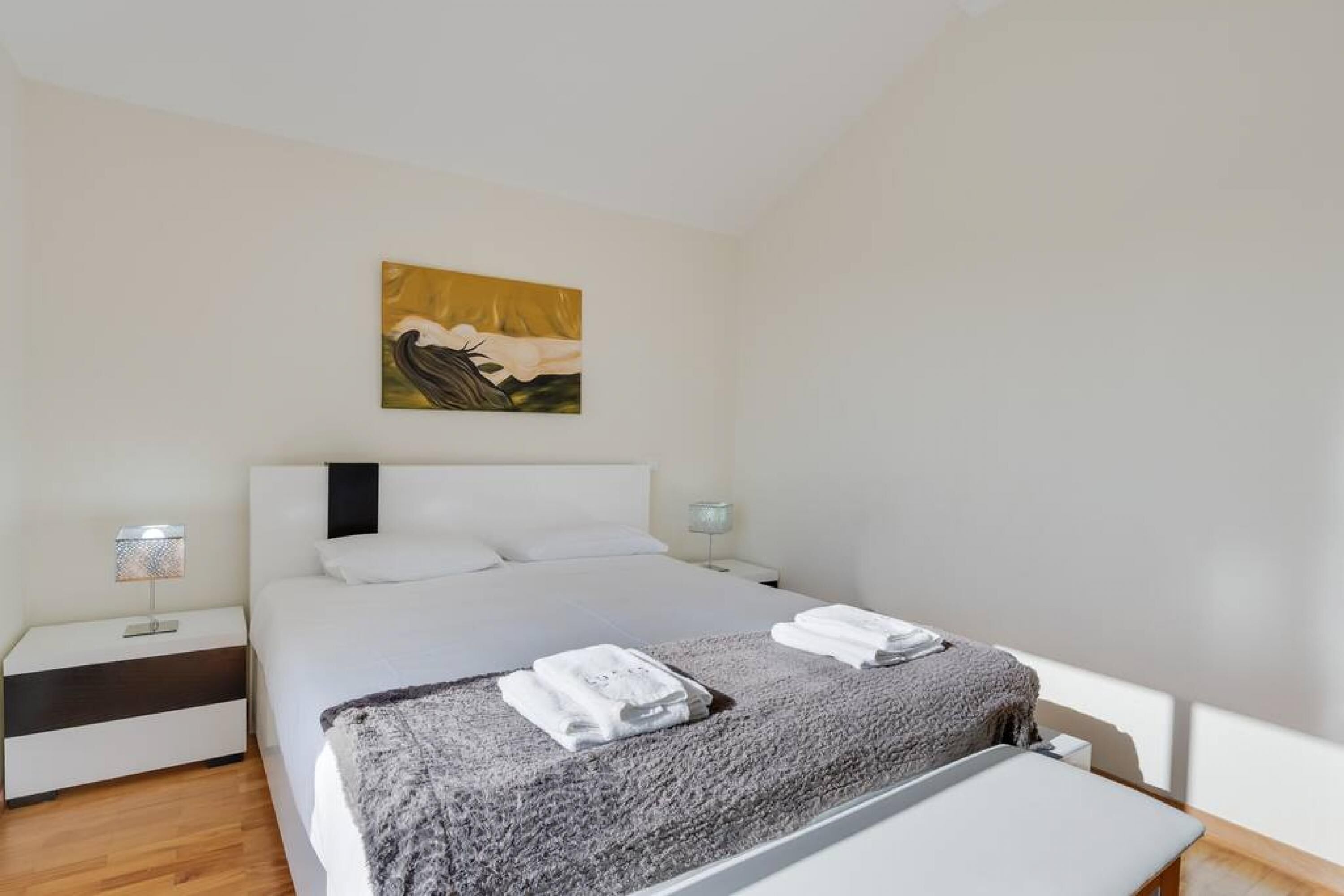 A hotel room with a large bed, two bedside tables, a nightstand, a painting on the wall, and a window providing natural light.