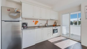 A modern kitchen with white cabinets, a stainless steel refrigerator, a built-in oven, and a large window offering a view of the outdoors. - Anje Apartamento 4 Q (Funchal)