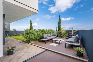Eine moderne Terrassenanlage mit einem grauen Sofa, zwei Couchtischen und einem kleinen Beistelltisch. Übertöpfe und eine Leiter lehnen an der Wand.