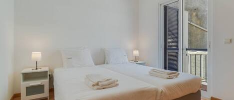 Cama doble con dos almohadas, mesita de noche con lámpara y ventana con persianas.