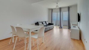 Ein modernes Wohnzimmer mit einem weißen Esstisch, vier Stühlen, einem grauen Sofa mit Kissen und einem kleinen weißen Beistelltisch. Ein Fernseher ist an der Wand montiert.