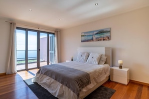 A bedroom with a large bed, a nightstand, a painting, and a view of the sea through a sliding door. - Canhas Views Pool and Barbecue (Ponta do Sol)