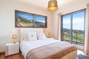 A bedroom with a large bed, two bedside tables with lamps, a large window with a view of a landscape, and a hanging light fixture. - Canhas Views Pool and Barbecue (Ponta do Sol)