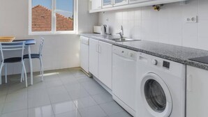 Et moderne køkken med hvide skabe, en indbygget vaskemaskine og et vindue med udsigt til en bygning.
