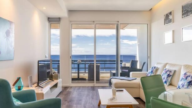 Una sala de estar moderna con un gran ventanal corredera que da a un balcón con vistas al mar.