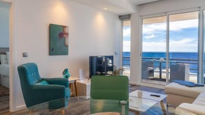 Una sala de estar moderna con una mesa de vidrio, sillas verde azulado y verde, un televisor de pantalla plana y un amplio ventanal con vistas al mar.