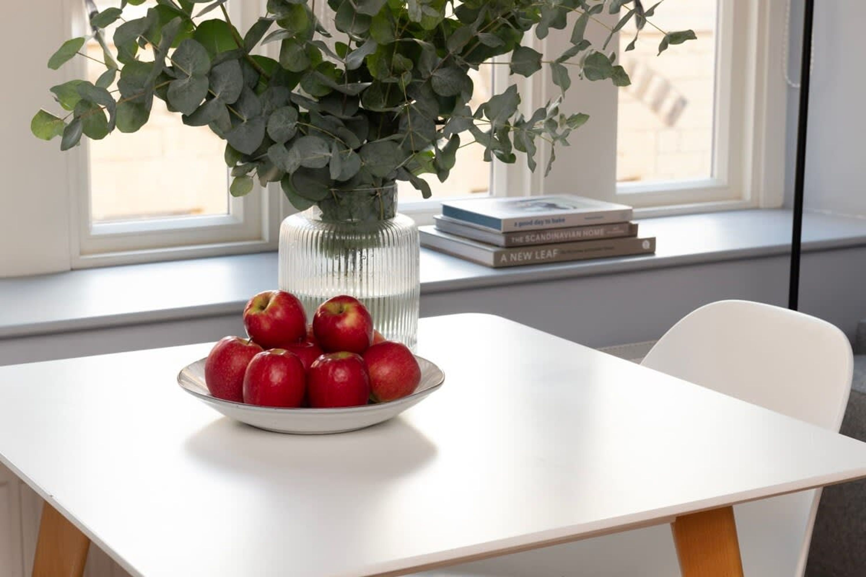 Una mesa blanca con un cuenco de manzanas rojas y un florero de hojas verdes.
