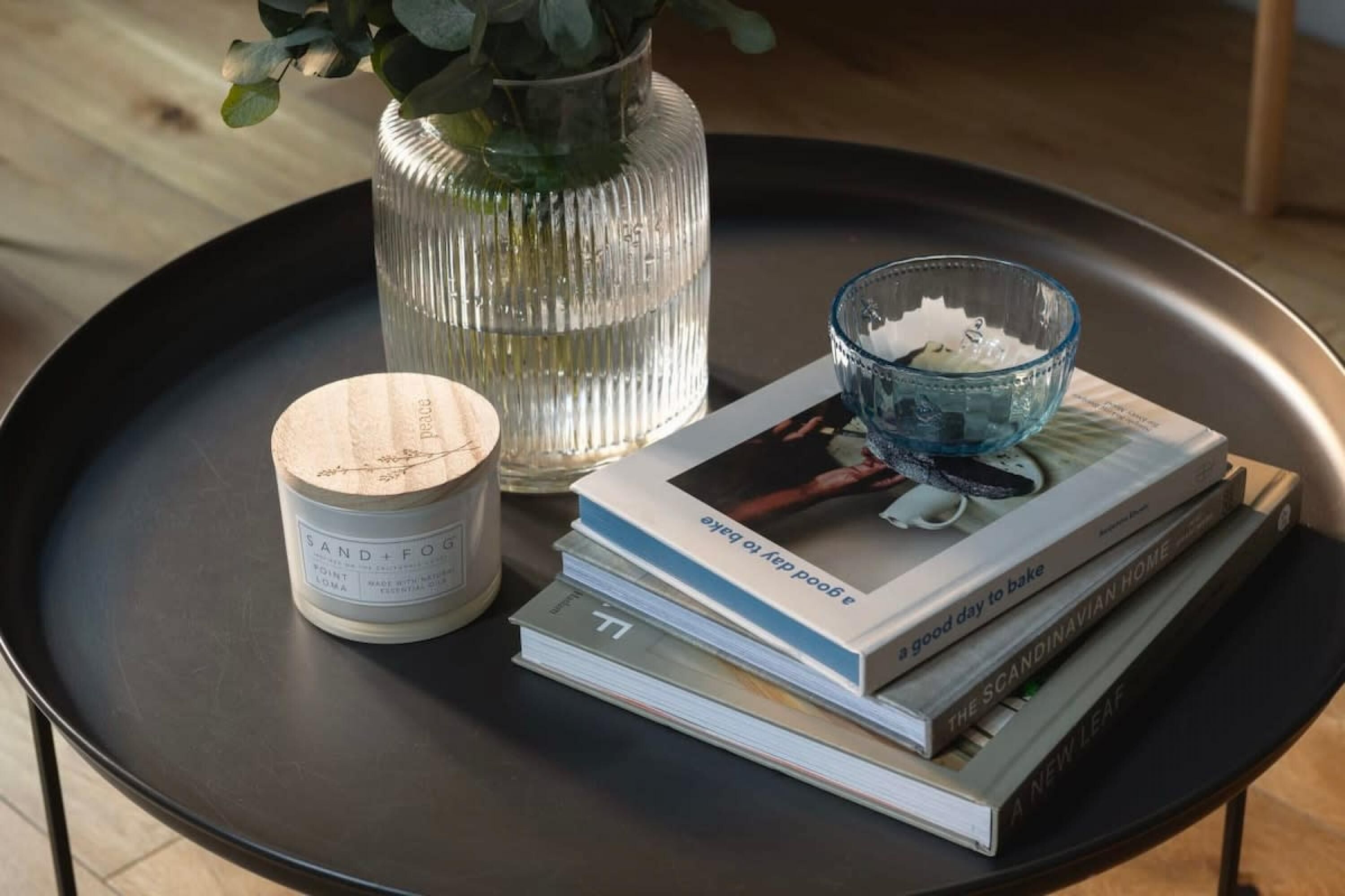 Une table ronde noire, une bougie, un vase en verre contenant des feuilles vertes et une pile de livres.