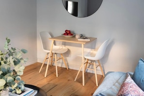 Un pequeño comedor con una mesa de madera y dos sillas blancas, un frutero y una taza sobre la mesa.