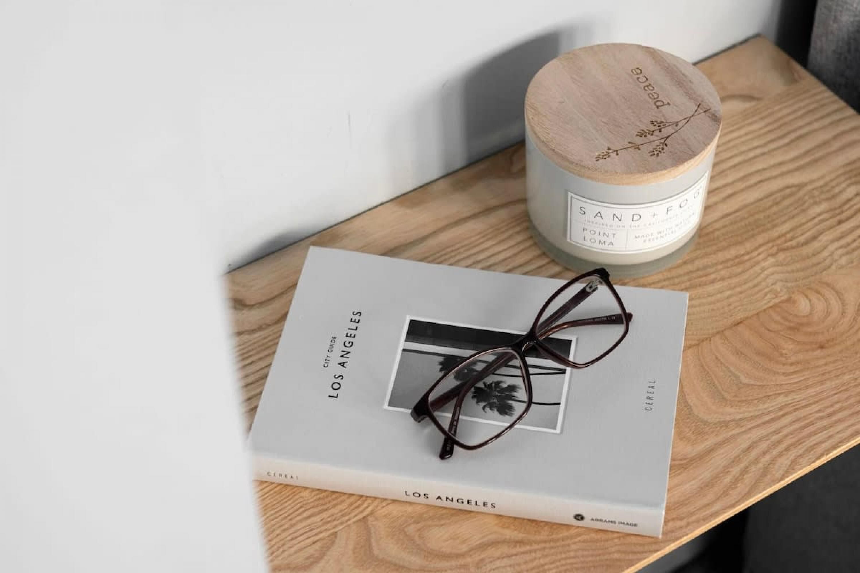 Un libro titulado "Los Angeles" con un par de gafas encima, sobre una superficie de madera junto a una vela etiquetada "Sand+Fox".