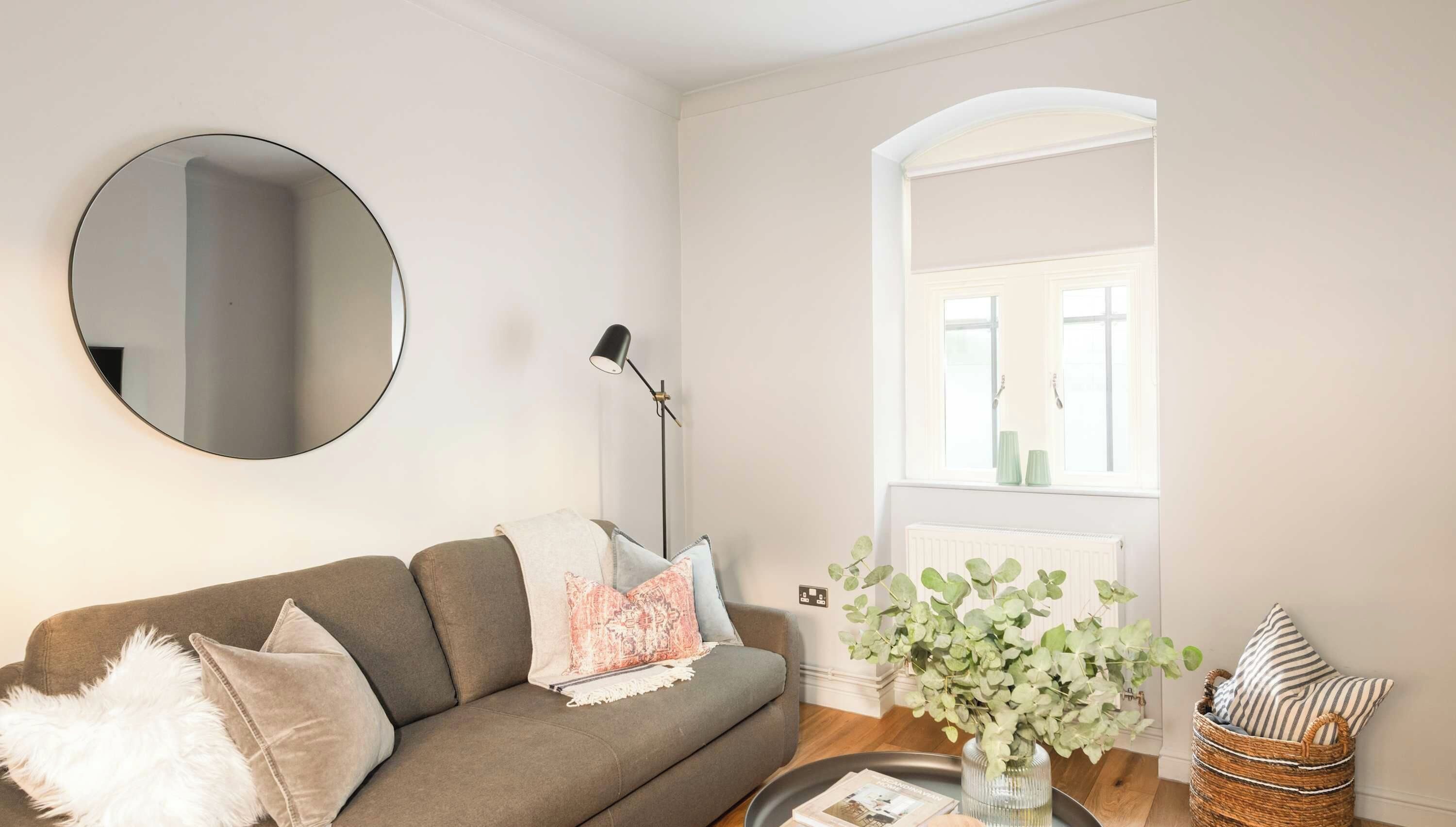 A living room with a sofa, a round mirror, a floor lamp, a window with a view, and a vase with plants.