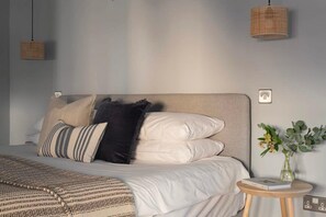 Ein ordentlich bezogenes Bett mit gemusterten Bettwaren und Kissen, ein kleiner runder Nachttisch mit einem Buch und einer Blumenvase, und eine gewebte Pendelleuchte darüber.