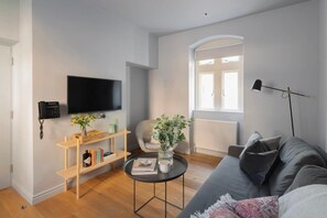 Ein modernes Wohnzimmer mit grauem Sofa, einem schwarzen Couchtisch, einem Bücherregal, einem an der Wand montierten Fernseher und einem Fenster mit Tageslicht.