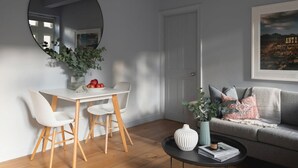 Una sala de estar moderna con una mesa de comedor blanca, dos sillas blancas, una mesa redonda de centro y un sofá gris. Un espejo redondo grande cuelga en la pared, y se exhibe un cuadro enmarcado sobre el sofá.