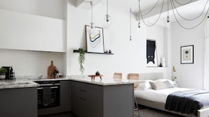 A modern kitchen with a built-in oven and dishwasher, a dining area with a bench and chairs, and a bed with a dark blanket. - Contemporary Studio - Minutes From Angel Tube St (London)