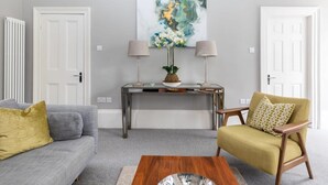Ein Wohnzimmer mit grauem Sofa, einem gelben Sessel, einem Couchtisch, einem Konsolentisch mit Lampen und einem großen abstrakten Gemälde an der Wand.