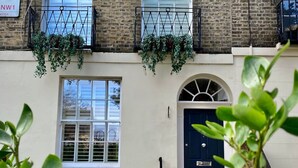 A two-story building with a brick facade, white window frames, and a dark blue door. There are two windows with black railings and hanging plants. - Luxury Marylebone House With Private Garden (London)