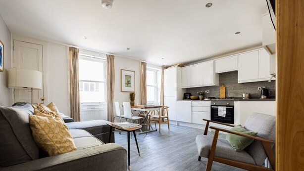 A modern living room with a sofa, armchair, and dining table. A kitchen with white cabinets and a stove is visible in the background. - Modern Duplex Near Oxford Street (London)