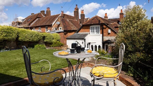 A patio with a table and chairs, a small garden, and a brick house with chimneys in the background. - Lavender Folly - Cosy Accommodation Alresford (Alresford)