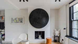 A modern living room with a large black textured wall art, a white chair, a small table with a candle, and a ceramic vase. - Stylish Georgian Townhouse w Sun Terrace Garden (London)
