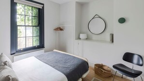 A bedroom with a bed, a window, a chair, a lamp, and a mirror on the wall. - Stylish Georgian Townhouse w Sun Terrace Garden (London)