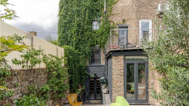 A brick building with a green ivy-covered wall, a balcony with a chair, and a small patio area with a table and chairs. - Stylish Georgian Townhouse w Sun Terrace Garden (London)