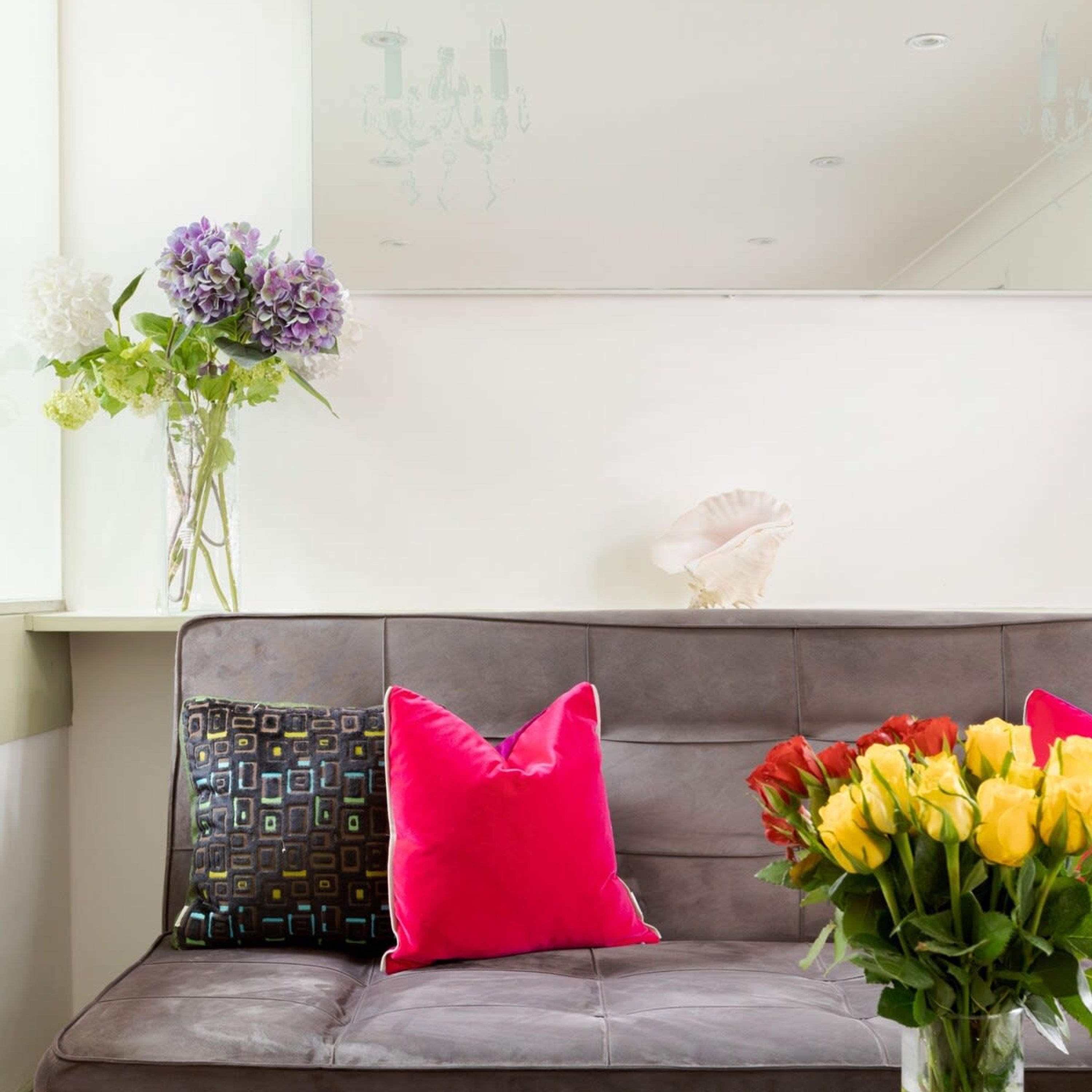A grey sofa with two pillows, one with a geometric pattern and one solid pink, and a vase with yellow and red flowers.