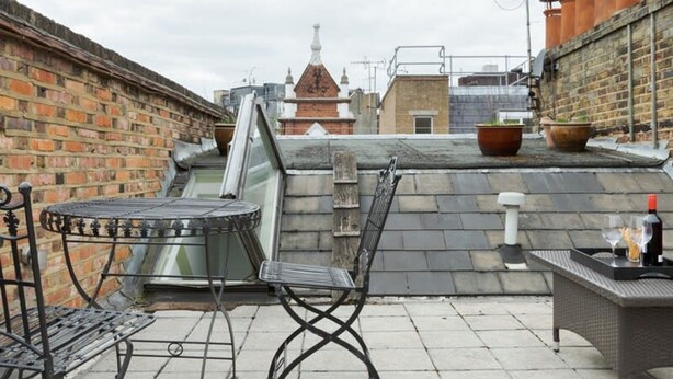 Une terrasse sur le toit avec une table et des chaises en métal, une petite table avec une bouteille et des verres, et un mur en briques.